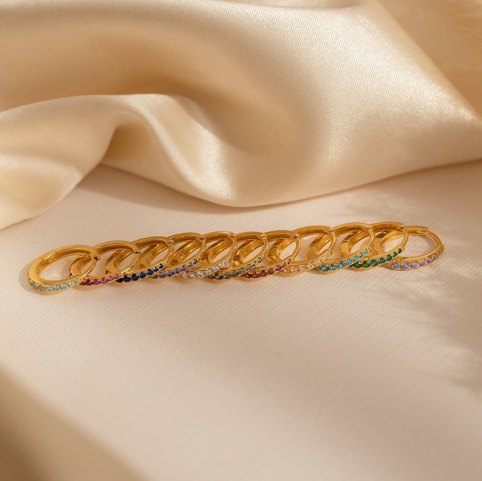 A row of gold rings, including Vera Pave Ruby Huggies with vibrant gemstones, displayed on beige satin fabric.