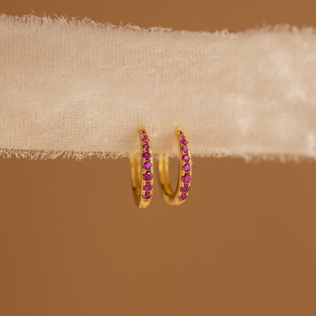 The Vera Pave Ruby Huggies are small gold earrings featuring pink stones, showcased on textured white fabric with a brown background.