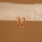 The Vera Pave Ruby Huggies are small gold earrings featuring pink stones, showcased on textured white fabric with a brown background.