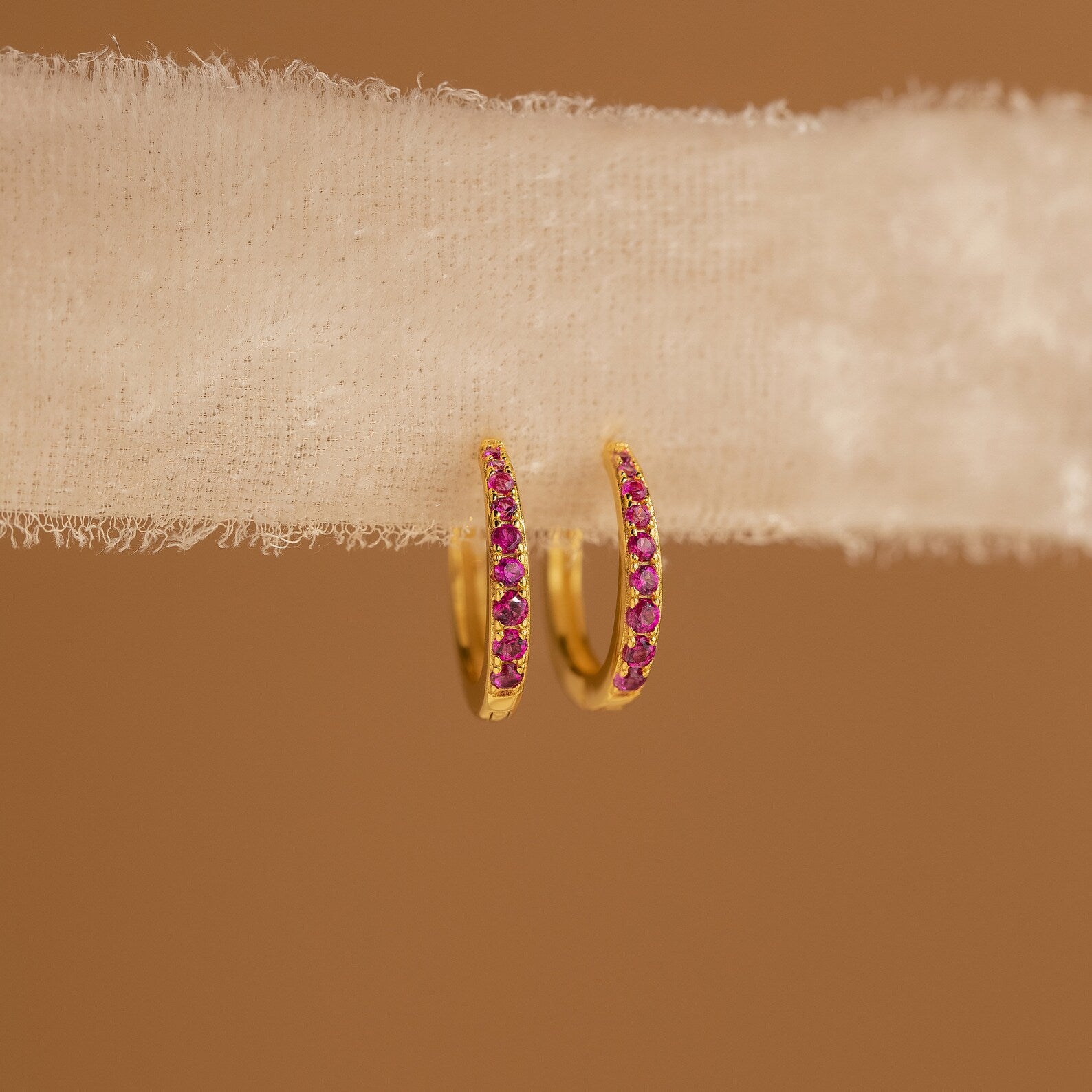 The Vera Pave Ruby Huggies are small gold earrings featuring pink stones, showcased on textured white fabric with a brown background.