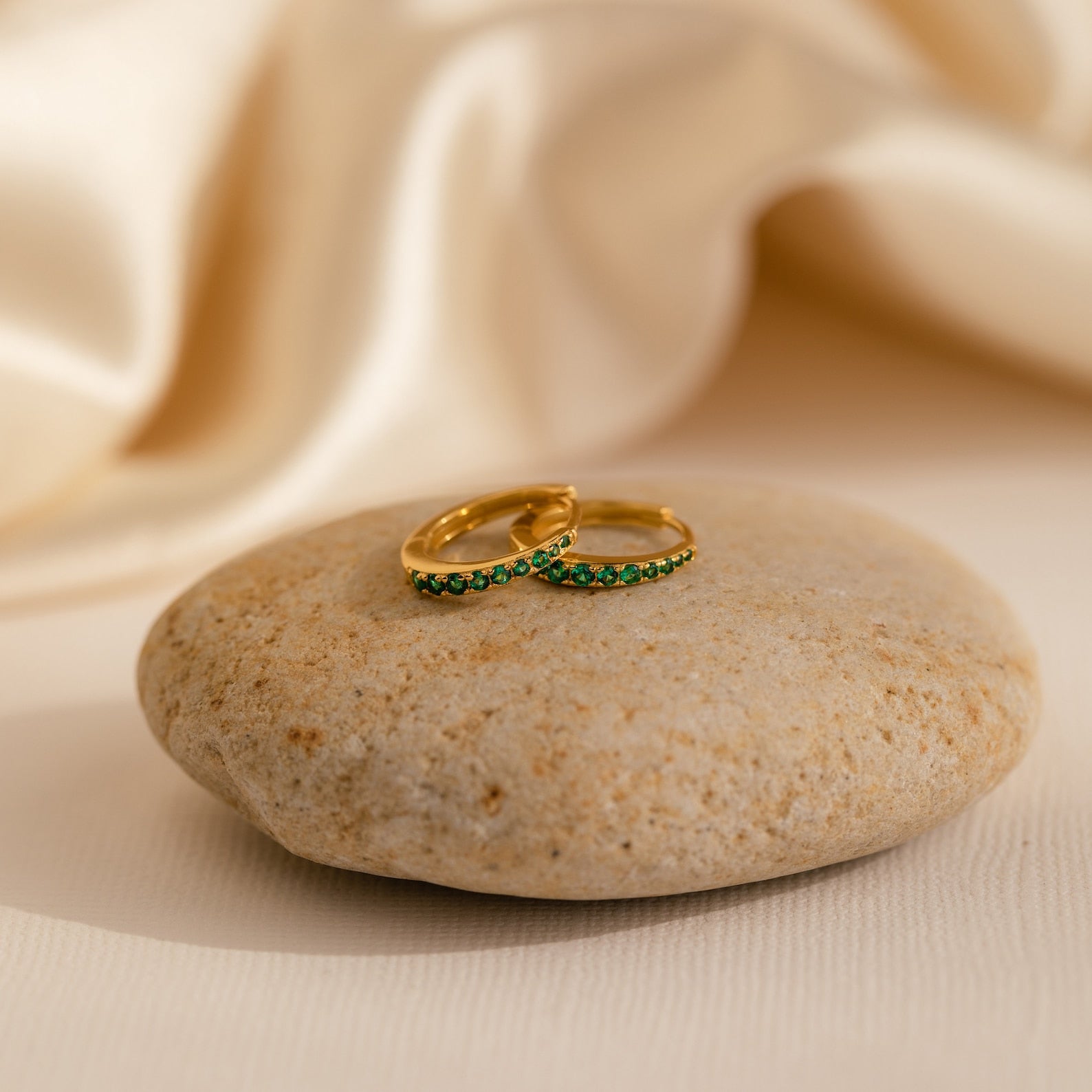 The Vera Pave Emerald Huggies, gold earrings with green stones, are displayed on a smooth stone against a soft beige fabric background.