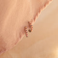 Pink Opal Huggies are gold dainty hoop earrings with small pink opal gems, displayed on soft peach fabric—perfect for adding a feminine touch to any look.