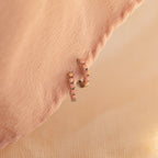 Pink Opal Huggies are gold dainty hoop earrings with small pink opal gems, displayed on soft peach fabric—perfect for adding a feminine touch to any look.