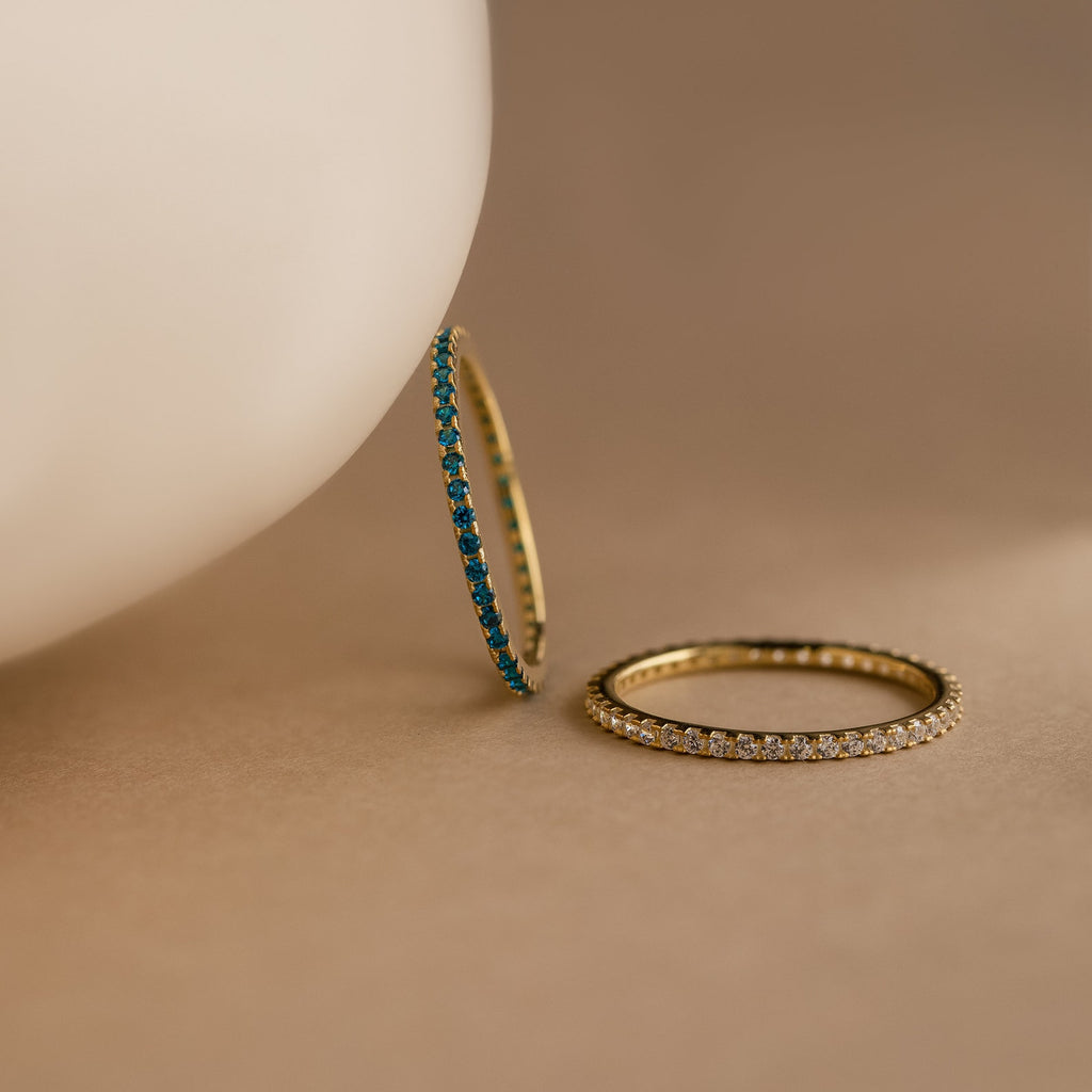 Two Eternity Birthstone Rings—one with blue gemstones, one with clear—rest on a beige surface. These customizable rings make a thoughtful Christmas gift.