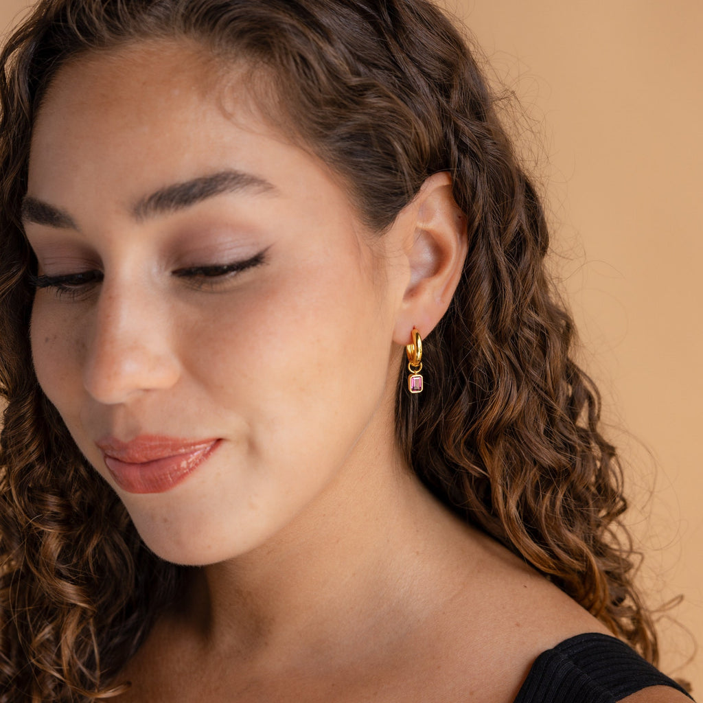 A woman with curly hair wears Dangling Gemstone Hoops with a gold finish and a black top, smiling softly against a beige background.