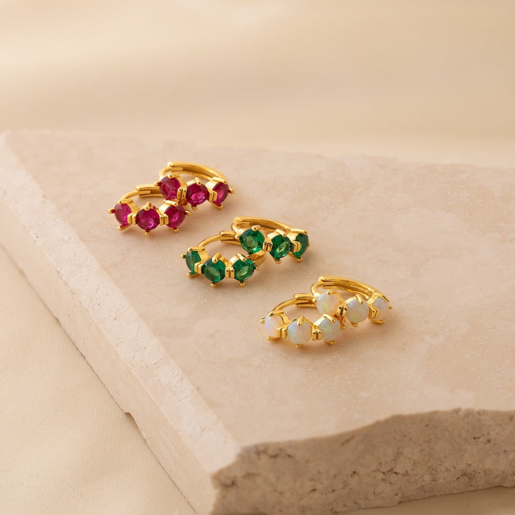 Three pairs of gold Triple Opal Huggies with pink, green, and white opal stones are displayed on a beige stone slab—perfect customizable minimalist hoop earrings.