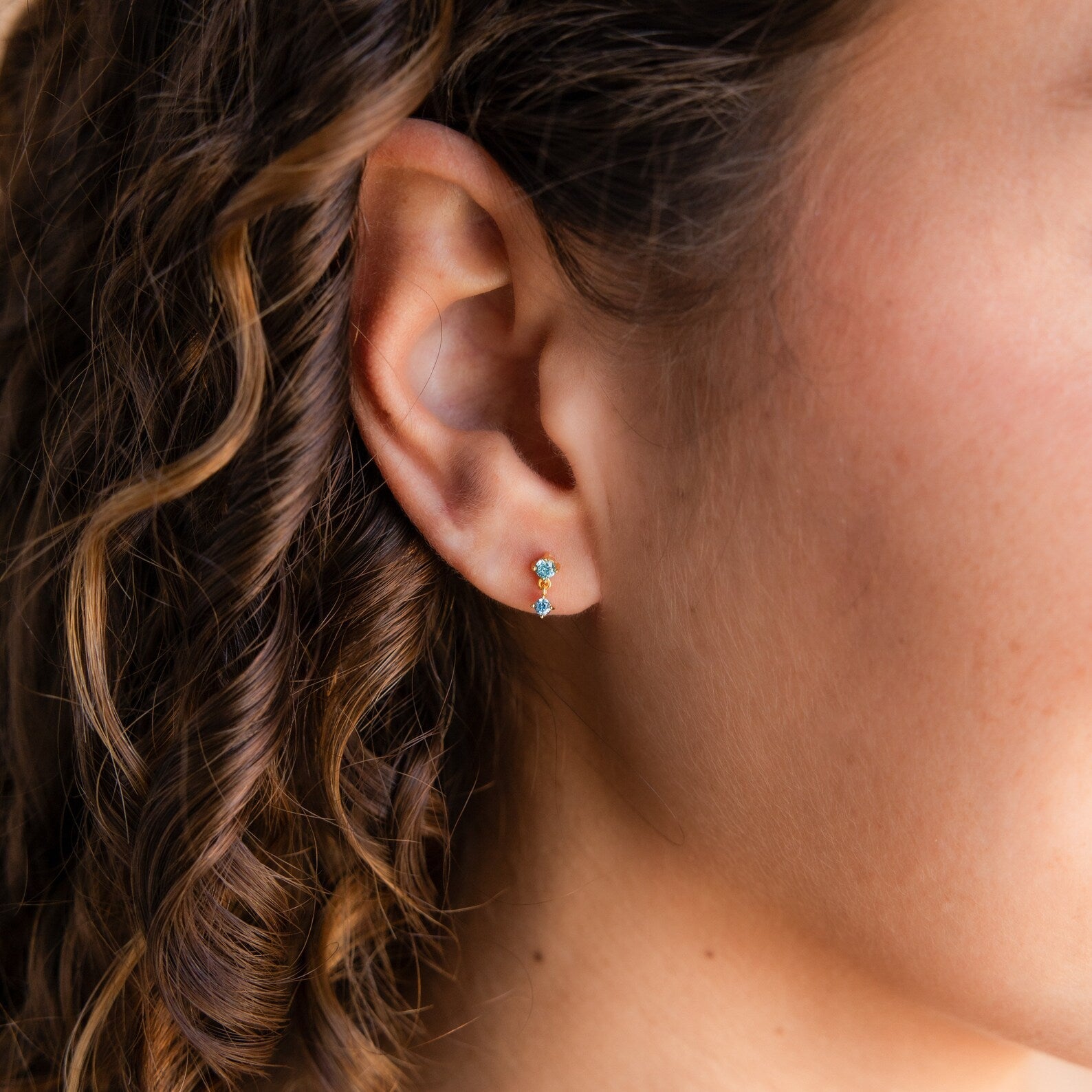Close-up of a woman's ear with an Amethyst Drop Stud—one of our elegant birthstone earrings—nestled amid curly brown hair framing her face.