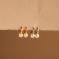 A pair of elegant Pearl Drop Huggie earrings is displayed against a textured, cream-colored paper backdrop. The design includes two sets of huggie hoops—one in shiny gold and the other in sleek silver—each holding a single lustrous round white pearl that dangles delicately beneath the hoop.