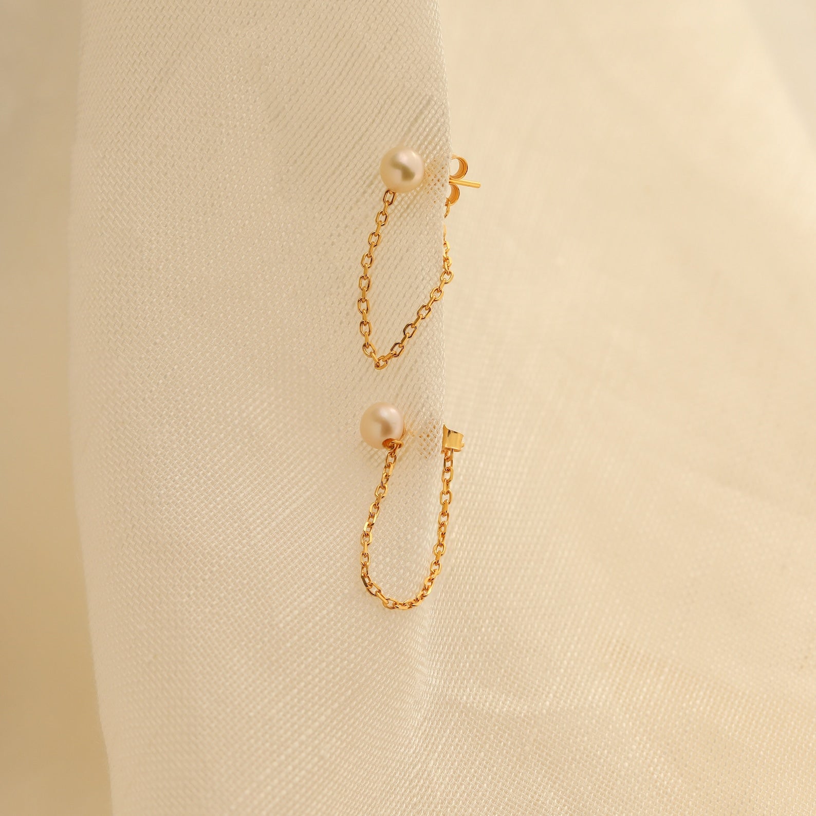 Pearl Chain Studs featuring elegant pearls on a delicate chain, showcased against a soft, cream-colored fabric background.