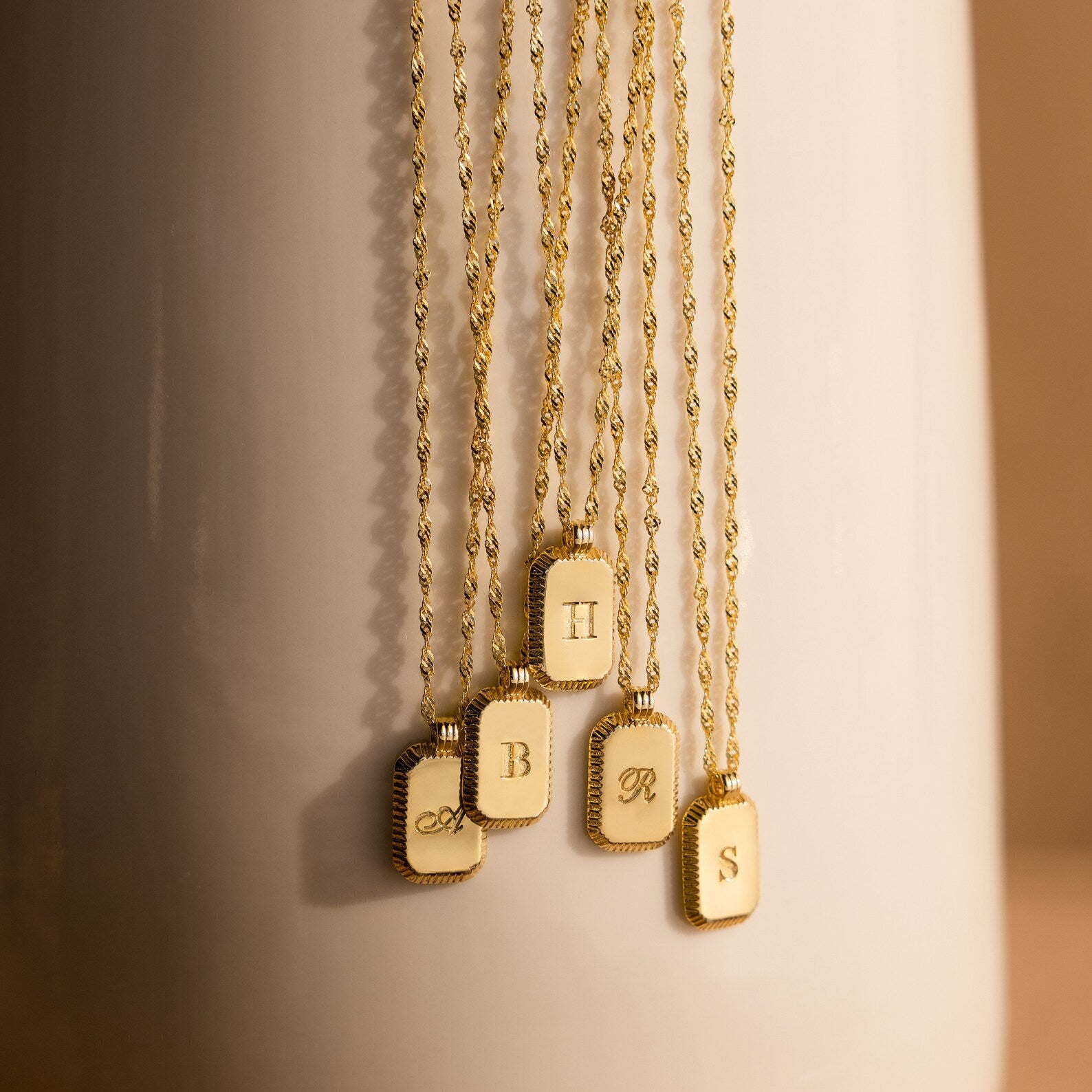 Newsletter Ribbed Initial Necklaces, featuring gold rectangular pendants engraved with A, B, H, R, or S, are elegantly draped on a vase for a stylish display.