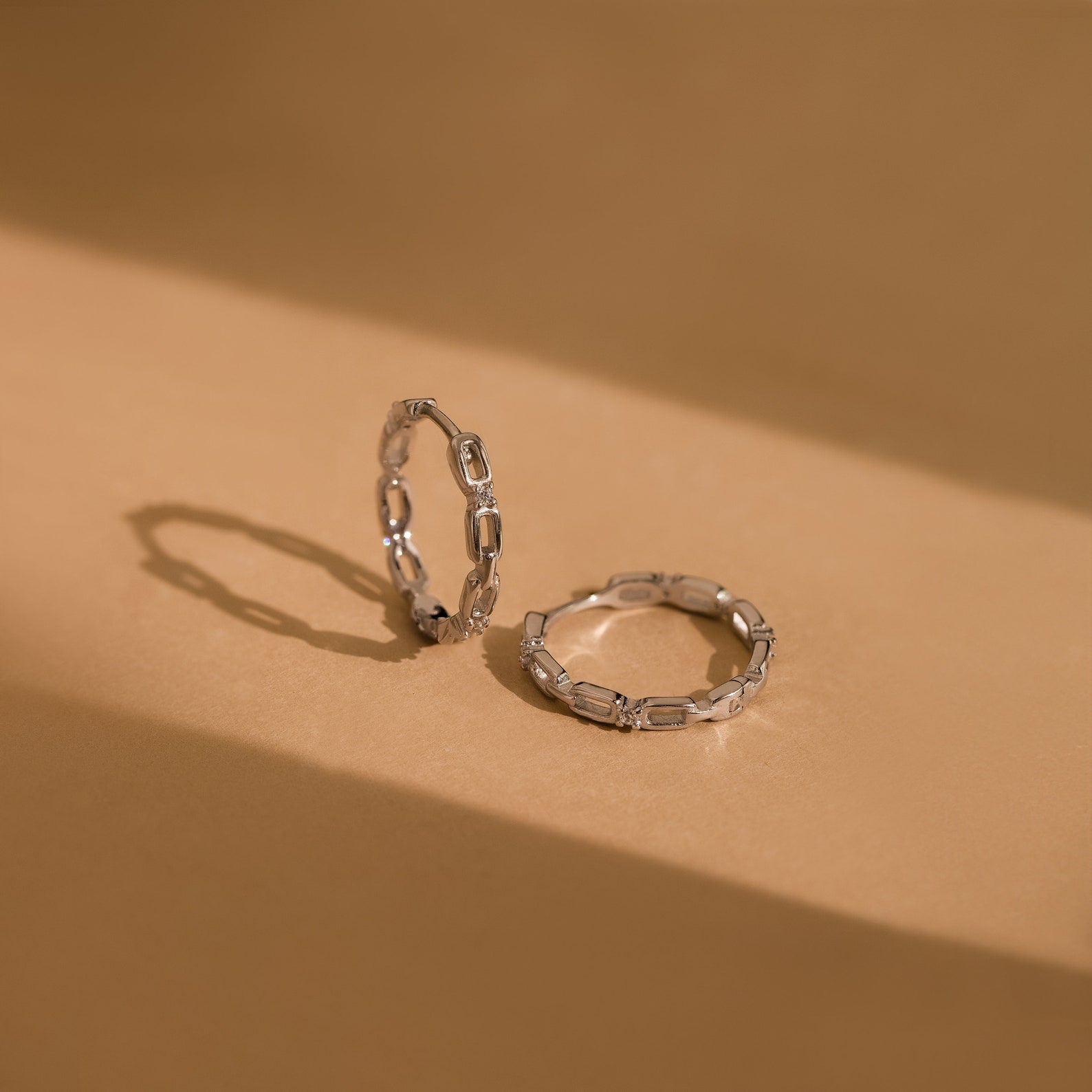 The Diamond Link Hoops—two silver chain-link rings on a beige surface with soft light—are perfect as bridal or wedding earrings, offering a subtle yet elegant touch.