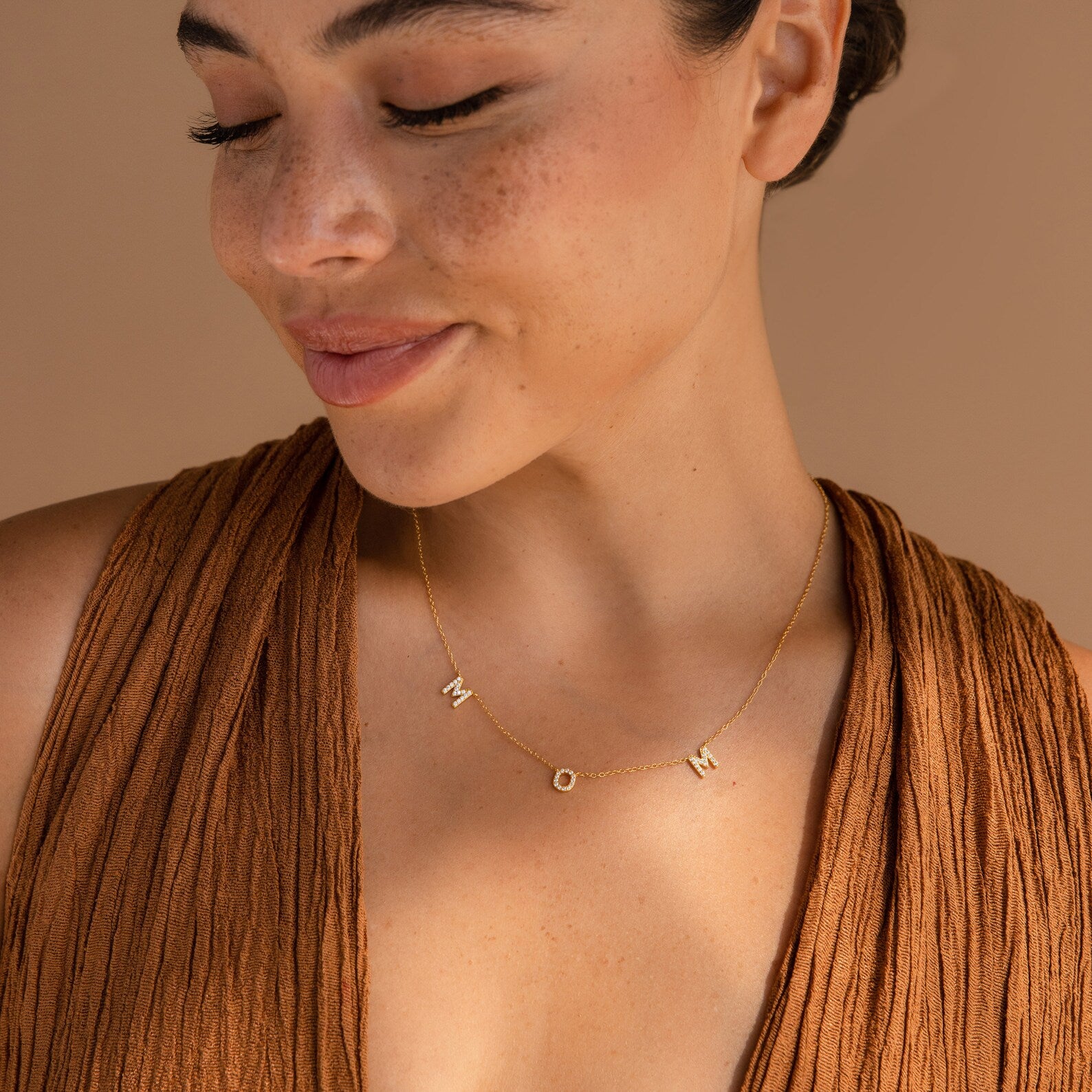 Smiling with her eyes closed in a brown top, she wears the Pave Mom Necklace—a heartfelt treasure with letter charms spelling MOM, perfect for gifting.