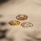 Three London Duo Name Rings in gold, rose gold, and silver are displayed on a beige fabric background.