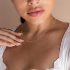 A woman showcases the Peach Station Necklace, pairing it with a white blouse and manicured nails gently resting on her collarbone for an effortlessly chic look.