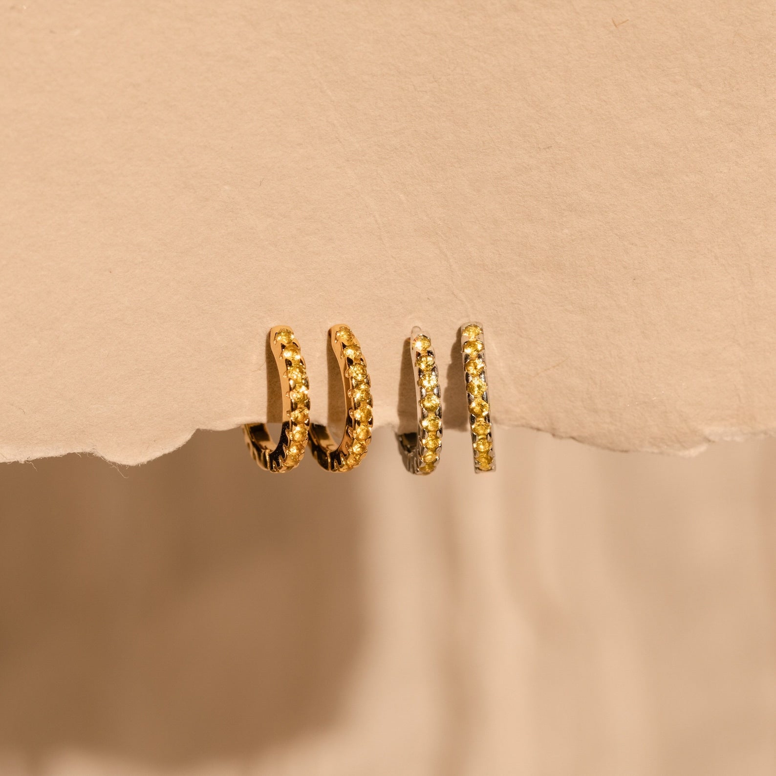 Three Citrine Pave Huggies hang elegantly on a torn piece of beige paper, their textured gold surfaces sparkling as they catch the light.
