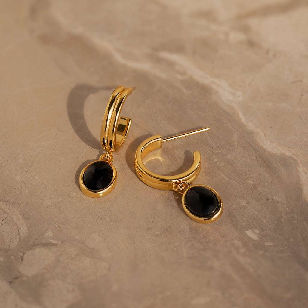 The Black Onyx Hoops Bundle, with striking black gemstone drops, is displayed elegantly on a beige marble surface.