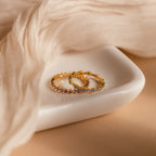 The Amethyst Eternity Ring and a minimalist gold ring rest on a white dish, with beige sheer fabric softly draped beside them.