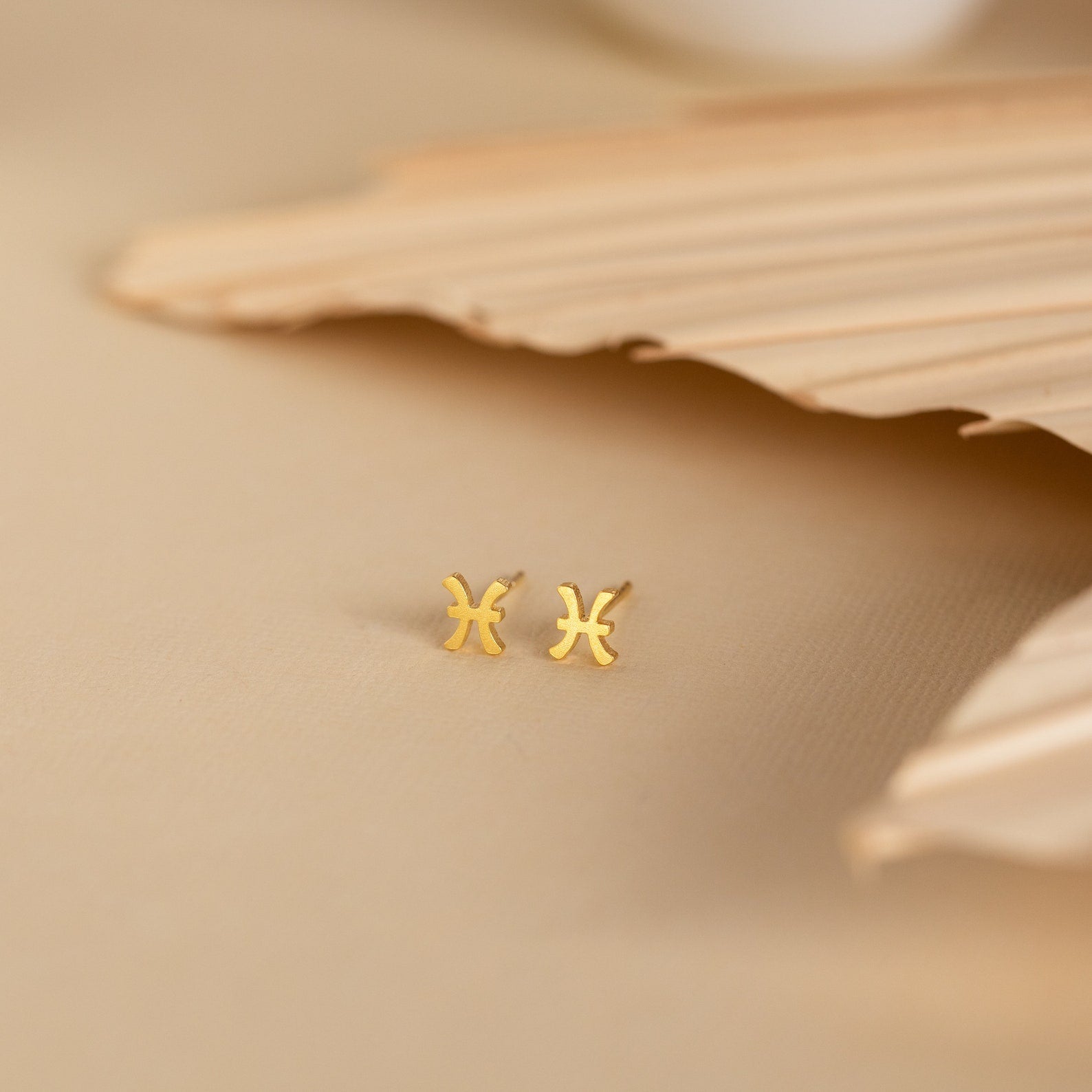 Tiny Zodiac Studs: small gold Pisces star sign earrings on a beige surface with textured leaf decoration in the background—perfect for adding a personalized touch to your earring stack.