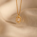 The Daydreamer Diamond Necklace features a gold pendant with a rectangular center stone framed by sparkling diamonds, exuding timeless elegance on a soft beige fabric background.