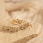 The Art Deco Ring Set features two delicate gold rings with clear gemstones, evoking vintage elegance, displayed on a textured stone surface against a soft beige background.