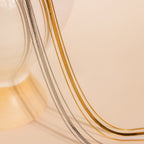The Triple Layered Chain Necklace in gold and silver is elegantly draped over the edge of a cream-colored vase, set against a beige backdrop.