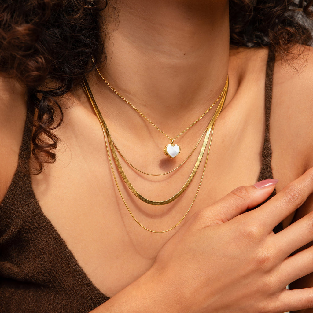 A woman wearing the Triple Layered Chain Necklace, which features gold herringbone and snake chains along with a heart-shaped pendant, rests her hand on her chest.