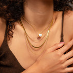 A woman wearing the Triple Layered Chain Necklace, which features gold herringbone and snake chains along with a heart-shaped pendant, rests her hand on her chest.