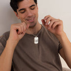 A smiling man in a brown shirt holds and admires a Men's Dog Tag Necklace with engraved design—making it the perfect Father's Day gift.