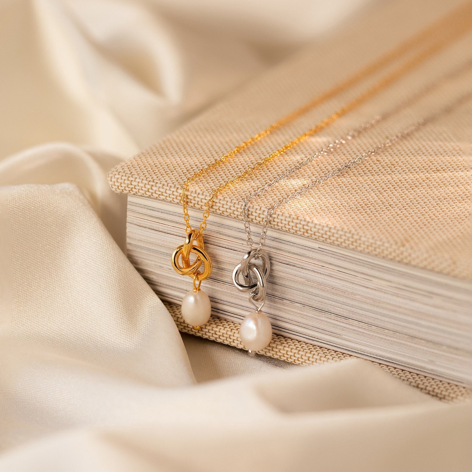 The Knot Pearl Necklace & Earrings Set in gold and silver, featuring pearl pendants, displayed on a textured book with cream fabric—the perfect gift for her.