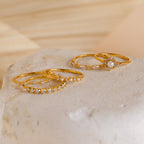 Three elegant gold rings from our Pearl Diamond Stacking Rings set, each set with small white gemstones, are showcased on a textured, light-toned stone surface.