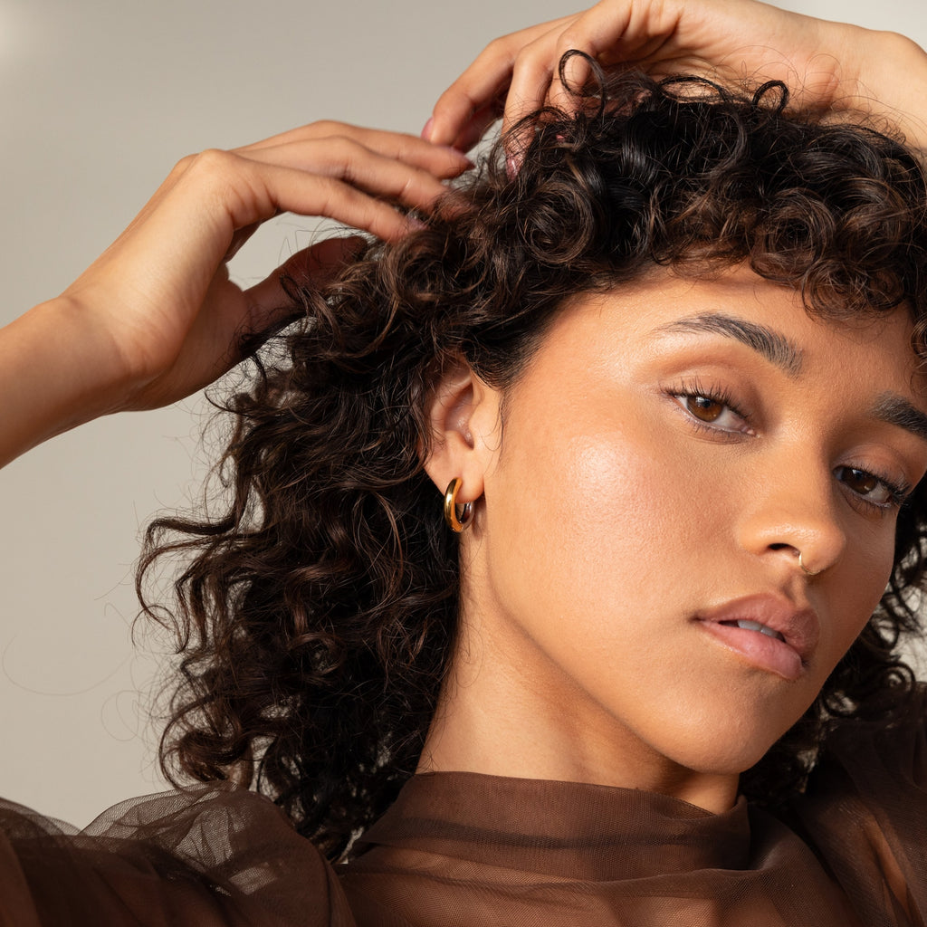 A woman with curly hair poses with her hands in her hair, wearing stylish Chunky Hoops.