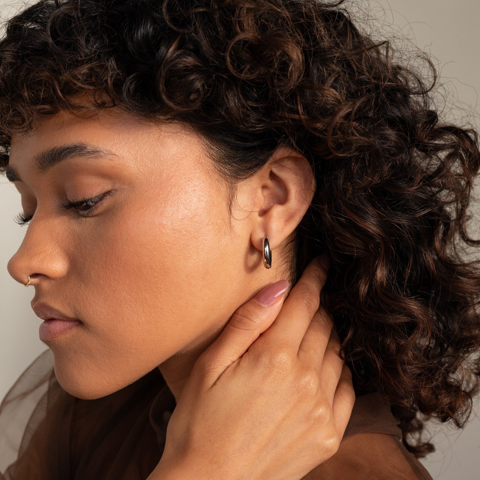 A person with curly hair touches their neck, highlighting smooth skin and wearing Chunky Hoops earrings.