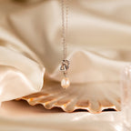 The Pearl Knot Necklace & Earrings Set in Sterling Silver features a delicate pendant displayed on a seashell against soft cream fabric—ideal elegant wedding jewelry.