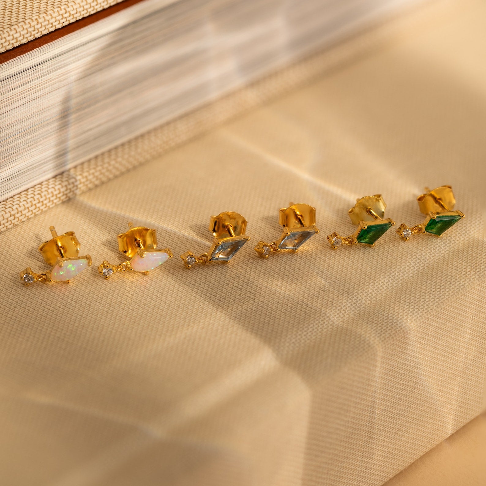 Three pairs of Art Deco Drop Studs—featuring opal, CZ diamond, and green stones—are displayed on a beige textured surface.