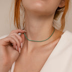 A woman with auburn hair in a white top wears the Emerald Tennis Choker Necklace, gently touching its elegant emerald gemstone as she highlights stylish necklace layering.