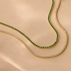 The Emerald Tennis Choker Necklace with vibrant green stones and a gold tennis necklace with clear stones are displayed on a beige surface—perfect for layering and adding style to any outfit.