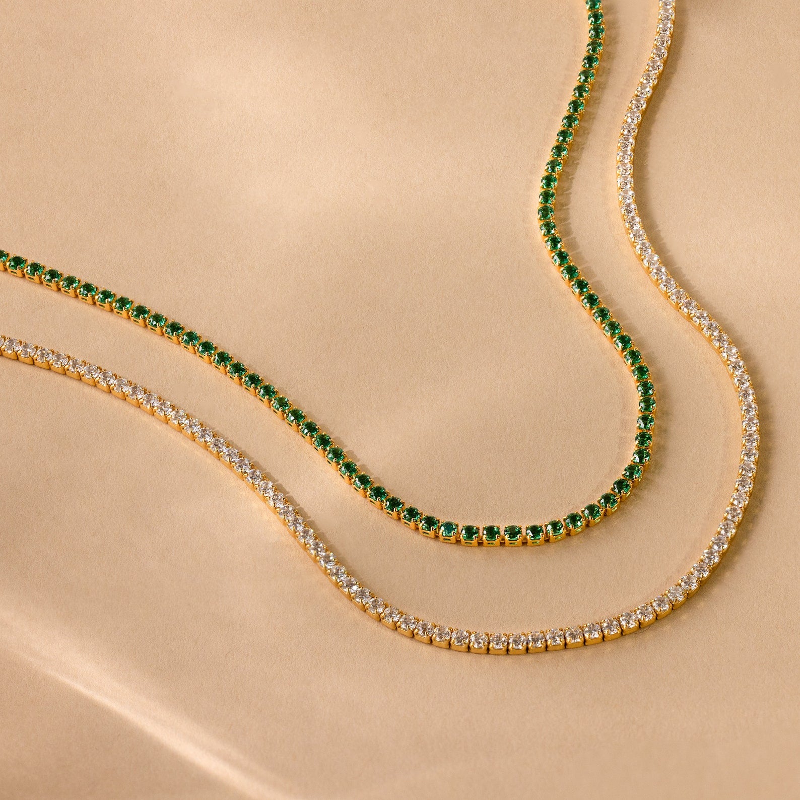 The Emerald Tennis Choker Necklace with vibrant green stones and a gold tennis necklace with clear stones are displayed on a beige surface—perfect for layering and adding style to any outfit.