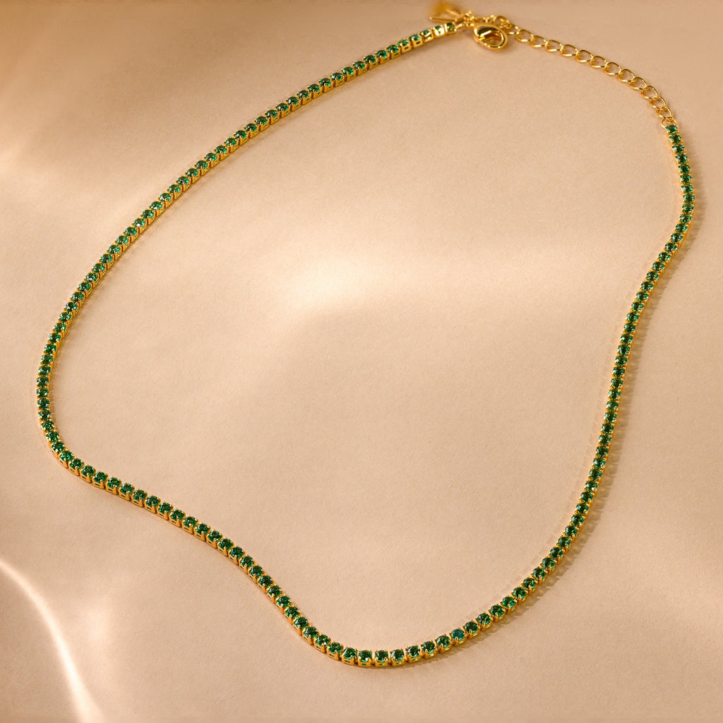 The Emerald Tennis Choker Necklace features a gold chain with delicate emerald gemstones, ideal for layering. Displayed on beige satin, this necklace adds a touch of elegance to any outfit.