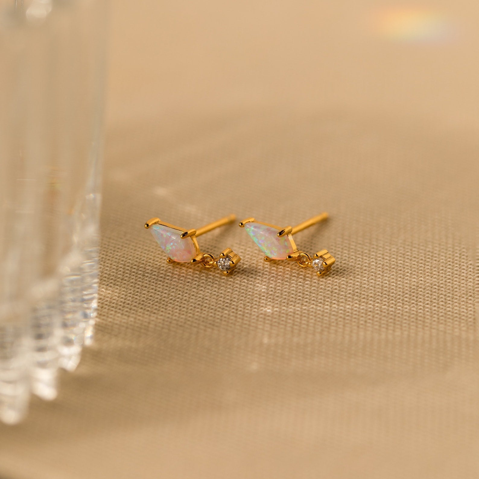 Opal Kite Drop Earrings with small crystal stones and CZ diamond accents rest on a beige textured surface near a glass edge, highlighting their elegant geometric design.