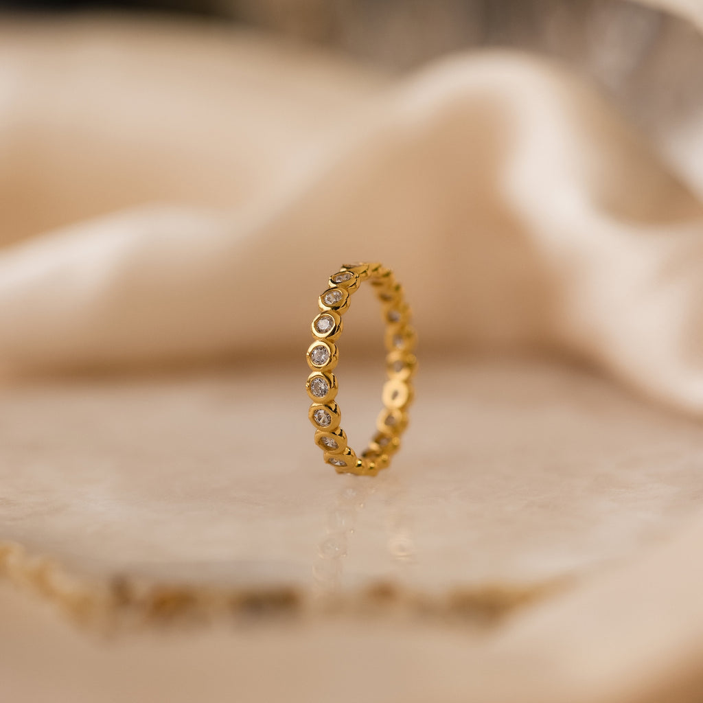 The Diamond Bezel Tennis Ring, featuring small round CZ gemstones, stands upright on marble with a soft beige background—ideal for stacking.