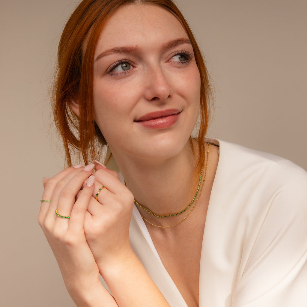 A red-haired woman in a white top smiles and looks to the side, wearing gold rings and layering necklaces, including the Emerald Tennis Choker Necklace.