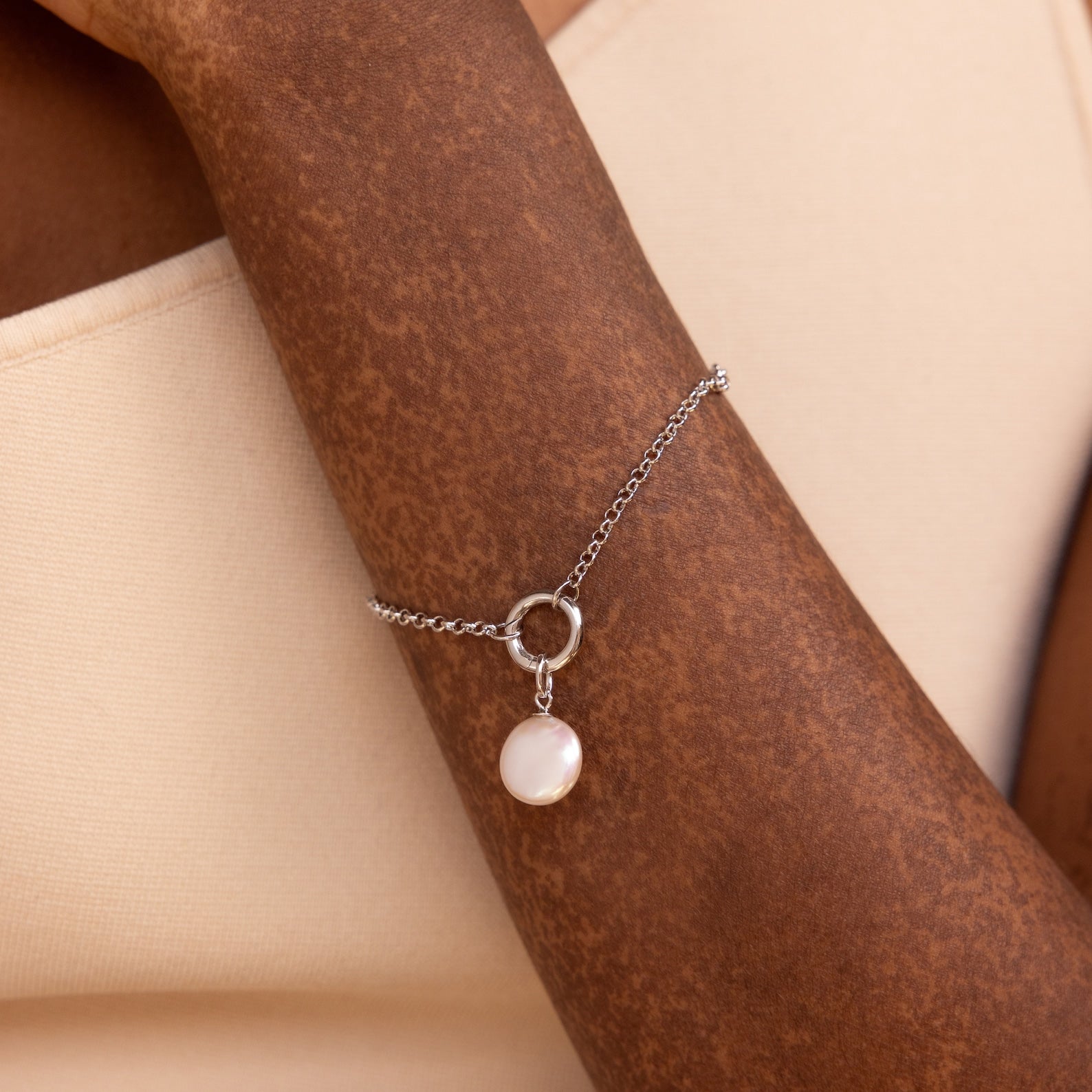 The Round Pearl Charm in Silver adorns a wrist with elegance, its delicate design standing out beautifully against beige fabric.
