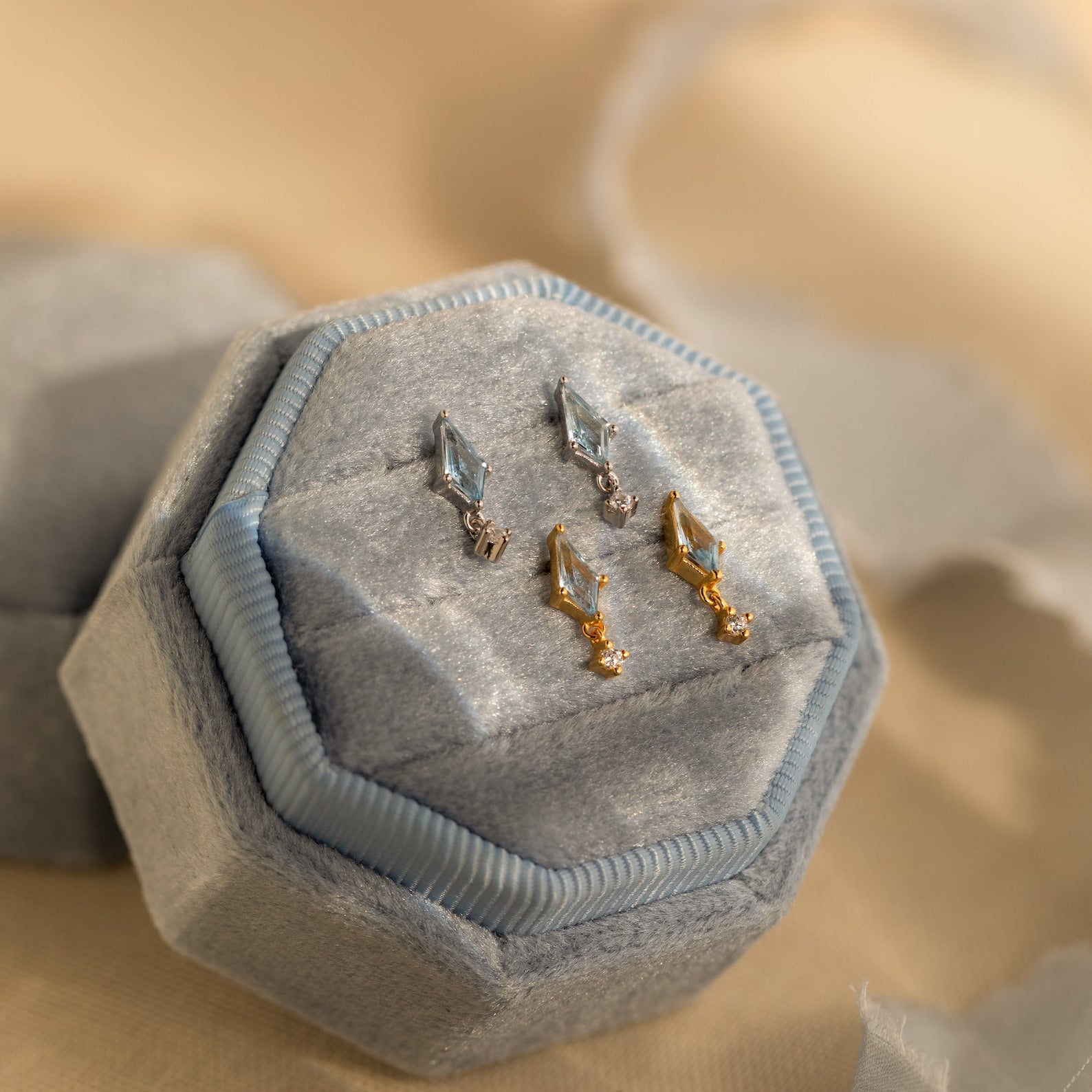 Two pairs of geometric earrings, including CZ Diamond and Aquamarine Kite Drop Earrings in silver and gold, displayed on a light blue velvet hexagonal jewelry box.