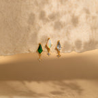 Three Opal Kite Drop Earrings, each with green, opal, and blue stones in a geometric design, are displayed on a textured beige surface under soft light.