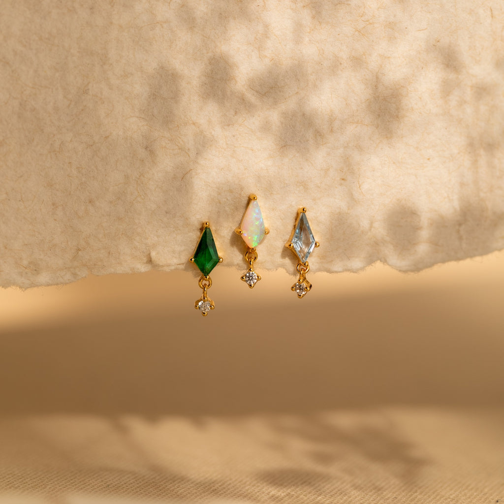 Three gold Aquamarine Kite Drop Earrings with green, white, and blue gemstones in a geometric design rest on a textured beige background with soft shadows.
