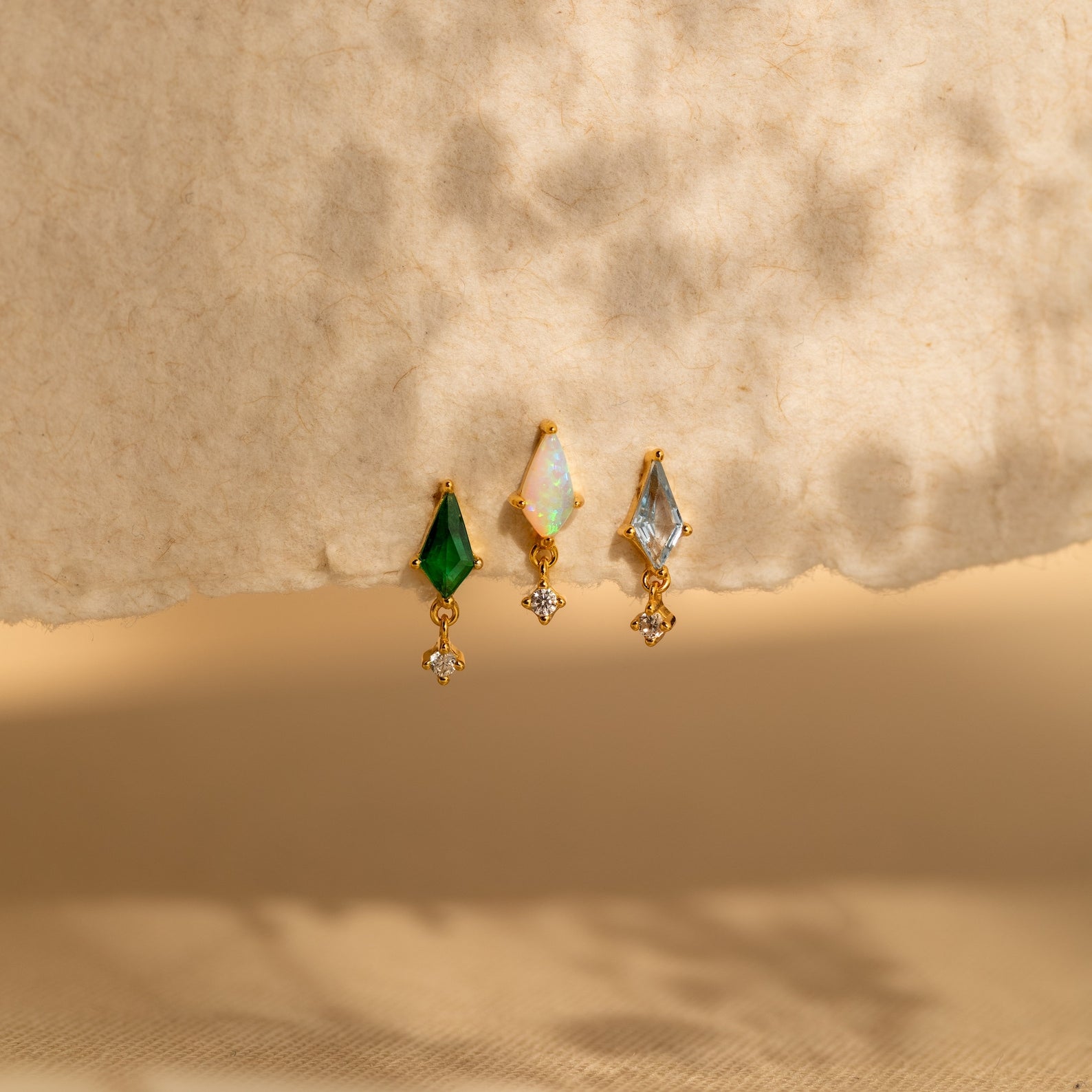 Three gold Aquamarine Kite Drop Earrings with green, white, and blue gemstones in a geometric design rest on a textured beige background with soft shadows.
