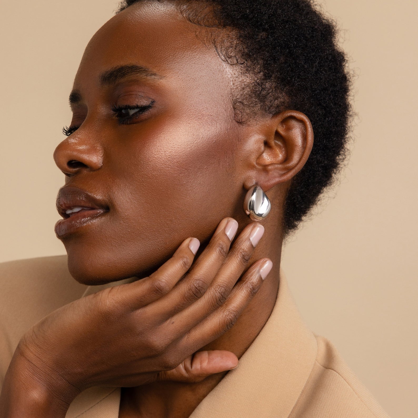 Woman with short curly hair wearing Bold Teardrop Studs, a beige blazer, and nude nail polish, posing thoughtfully.