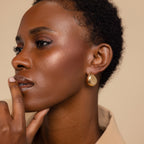 A woman with short curly hair wears Bold Teardrop Studs and touches her lips, gazing thoughtfully to the side.