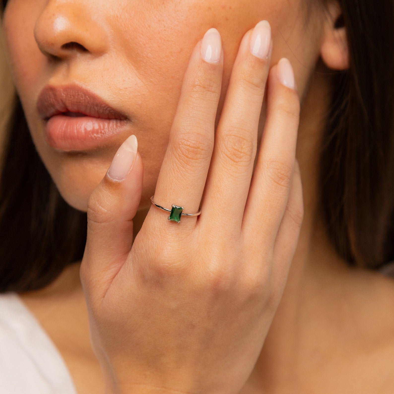 A woman touches her face, wearing the Harley Emerald Ring - Silver—a vintage-style ring featuring a small green gemstone on her finger.