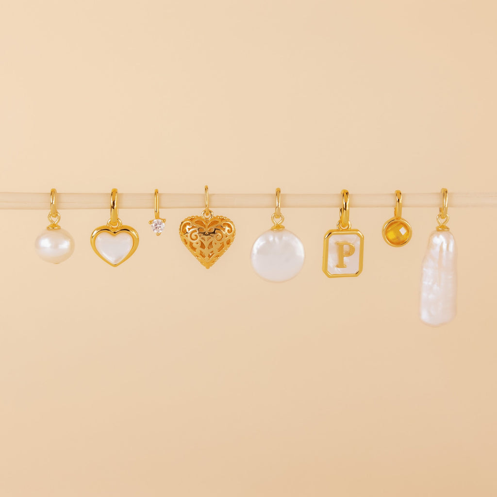 Eight gold and pearl charms, including the Round Pearl Charm in 18K Gold and a heart-shaped Mother of Pearl pendant, are displayed on a beige background.
